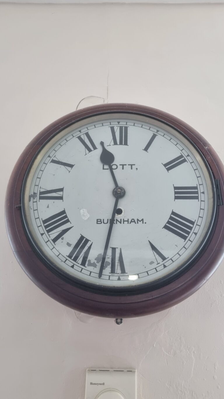 Round wall clock with a burgundy frame and Roman numeral dial on a pale wall, showing a worn white face.