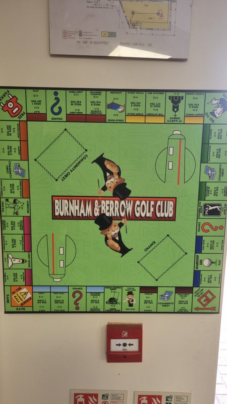 Giant green Burnham & Berrow Golf Club Monopoly board poster on a wall, with a red fire alarm box mounted below it.