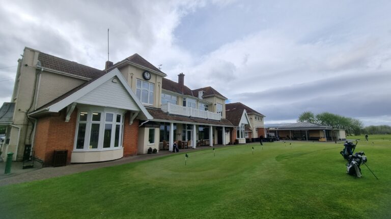 Two-story clubhouse with clock, beige walls, white trim, and a red-tiled roof beside a manicured golf green.