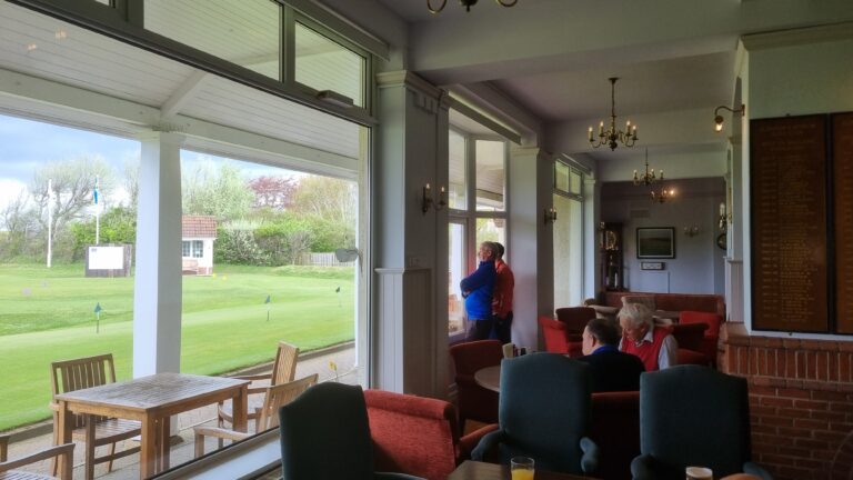 Indoor golf club lounge with large windows looking out onto a green lawn; two men stand by the window while others sit on red and blue chairs and sofas under chandeliers.