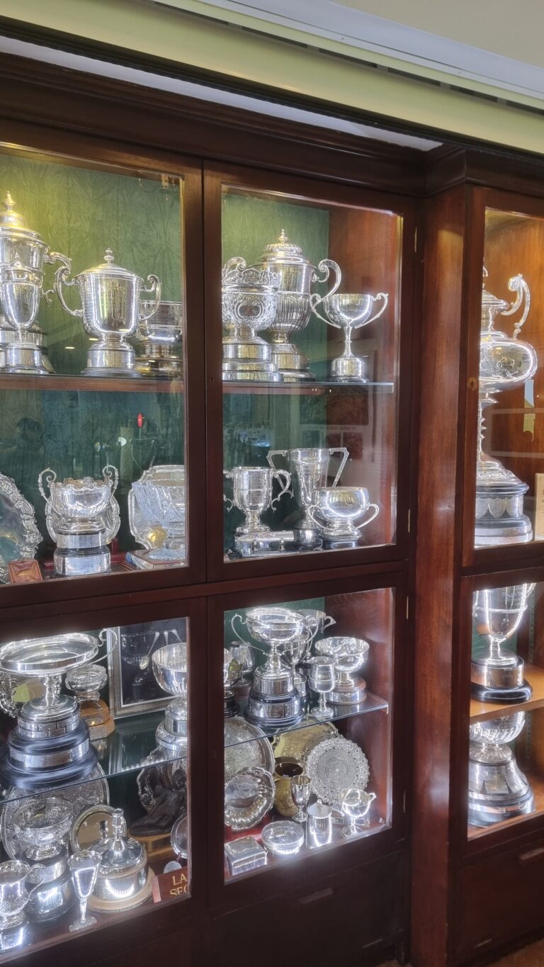 Wooden display cabinet with glass doors showcasing silver trophies and cupware inside.
