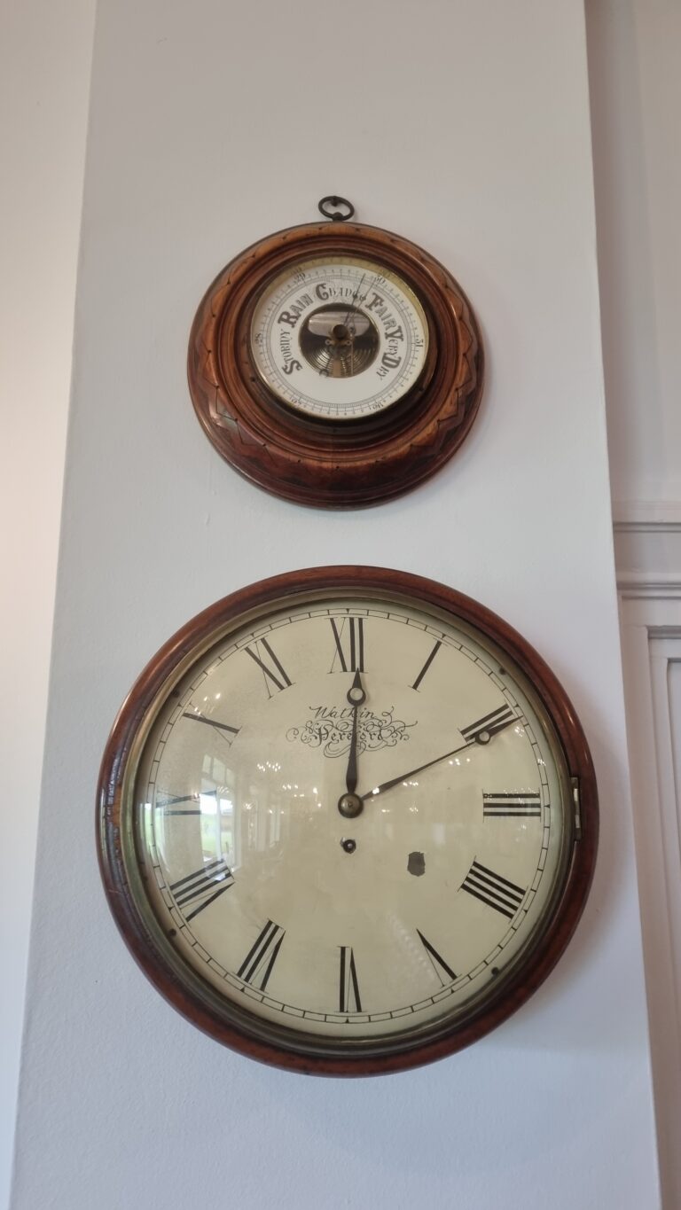 Two antique round wall clocks mounted on a light wall: a smaller top clock with a wooden frame and inner dial, and a larger bottom clock with Roman numerals.