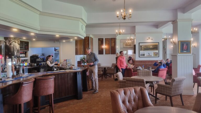 Pub interior with a long bar, back counter, and brown leather stools; patrons seated at tables under chandeliers.