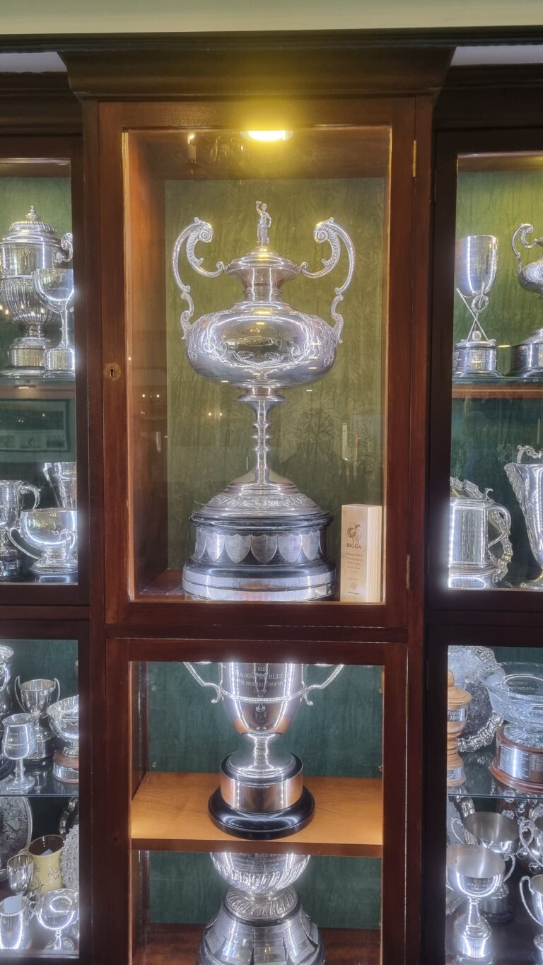 Silver ornate trophy displayed in a wooden glass cabinet, center shelf, with other trophies visible around it.