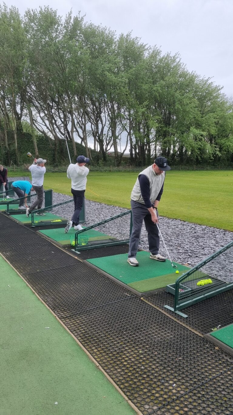 People at a golf driving range practicing swings on padded mats outdoors with trees in the background.
