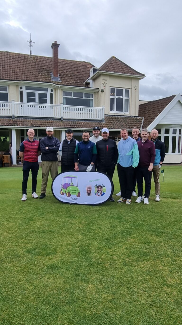 Group of men posing on a golf course with a banner in front, smiling faces and a large house in the background.