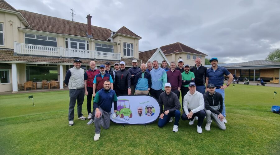 Group of male golfers posing for a photo on a putting green, with a banner and clubhouse in the background on a cloudy day.