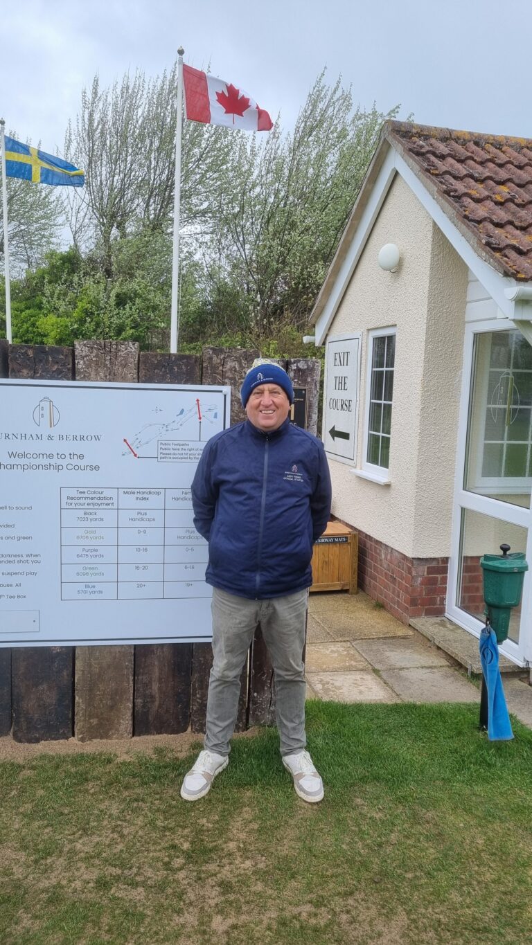 Man in a blue jacket and beanie standing beside a course sign and a large information board outside a building with flags in the background (Canada and Sweden).