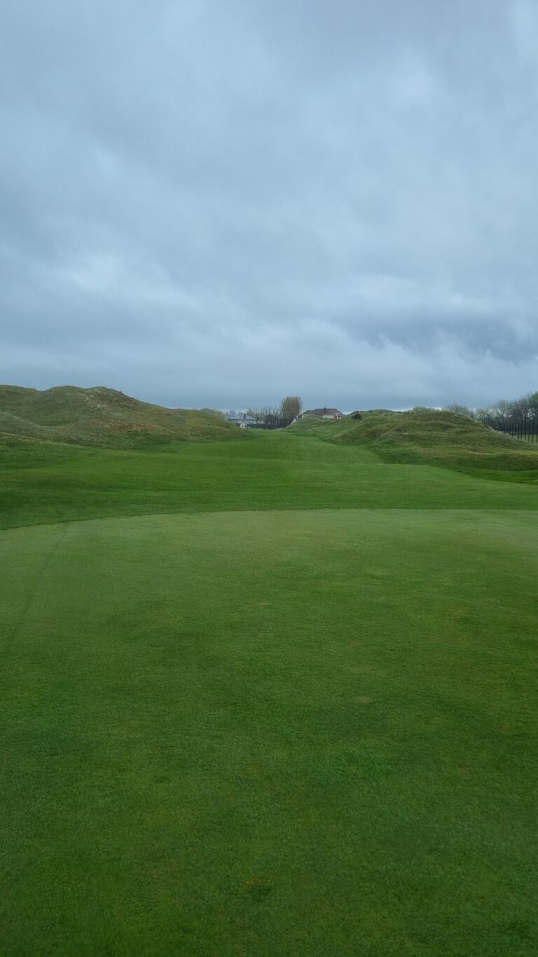 Green golf fairway with rolling mounds and distant houses under a cloudy blue sky