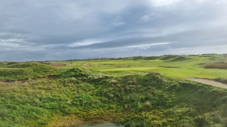 Open golf course panorama with green fairways, sand dunes, and a cloudy blue-gray sky in the distance.