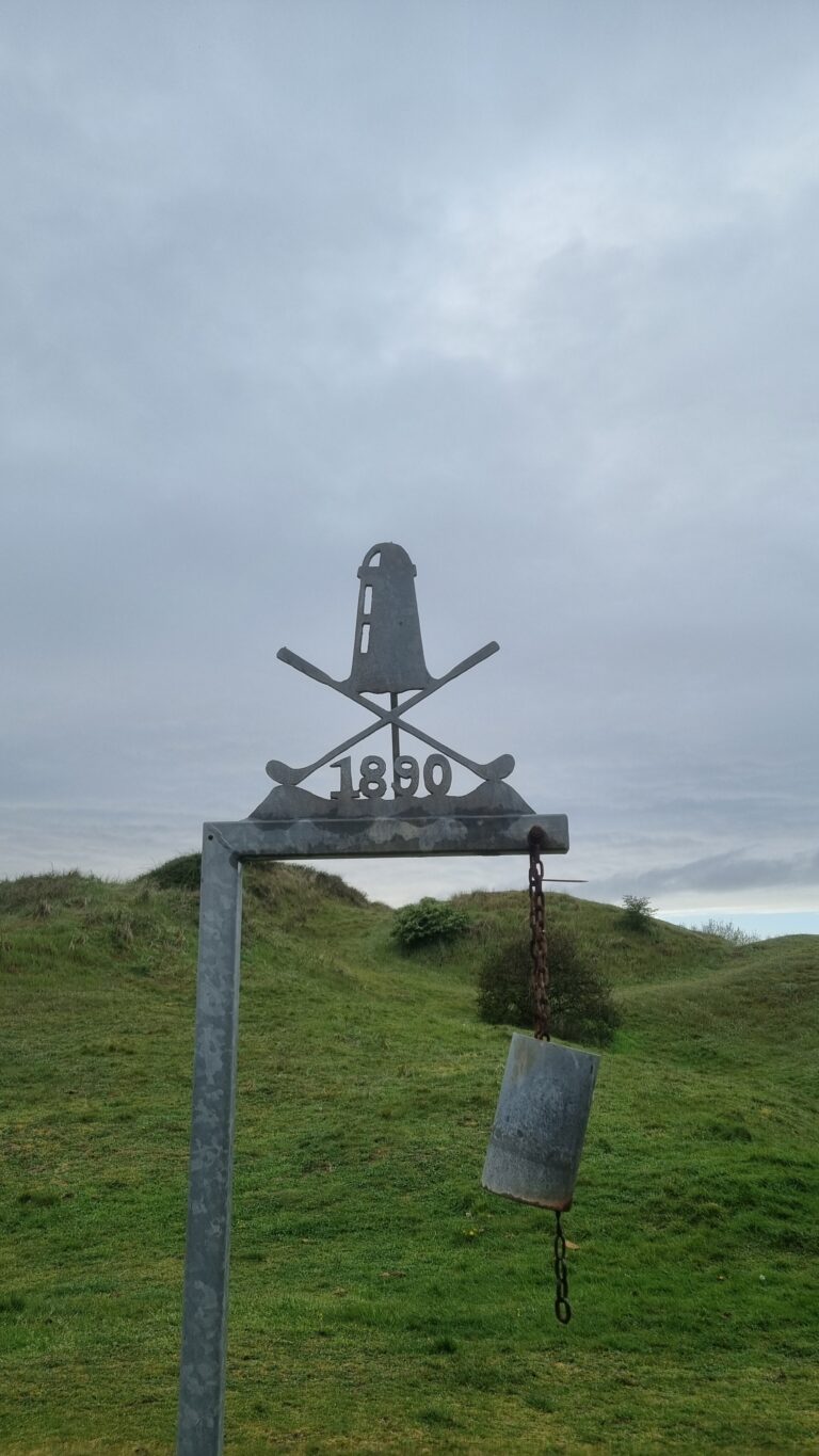 Outdoor metal monument on a frame, displaying '1890' with two crossed pickaxes and a vertical emblem, chain and padlock hanging from the frame over a grassy hill.