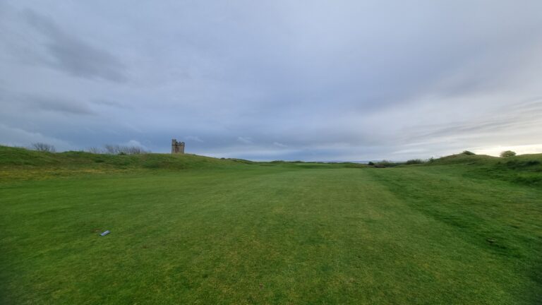Wide green field with a small stone tower on a distant hill under a cloudy sky