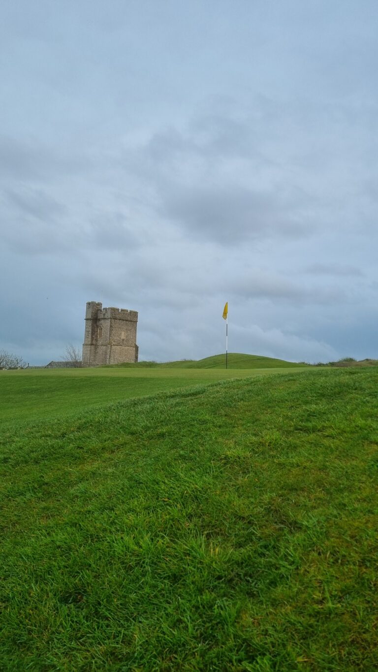 Green grassy hill with a small stone tower on the left and a yellow flag on a pole on the right under a cloudy sky.