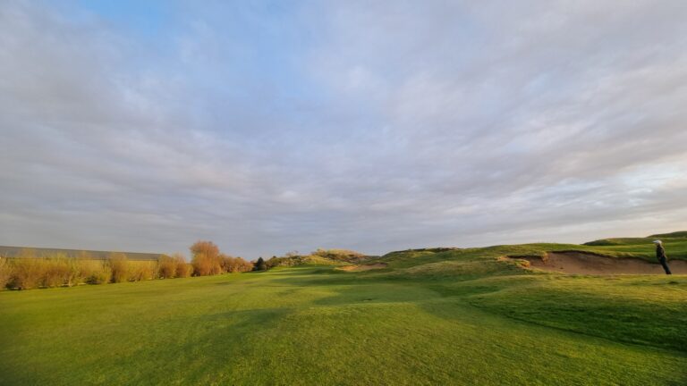 Expansive golf course with a green fairway, a sand bunker on the right, and a person wearing a hat standing on a path under a cloudy sky.