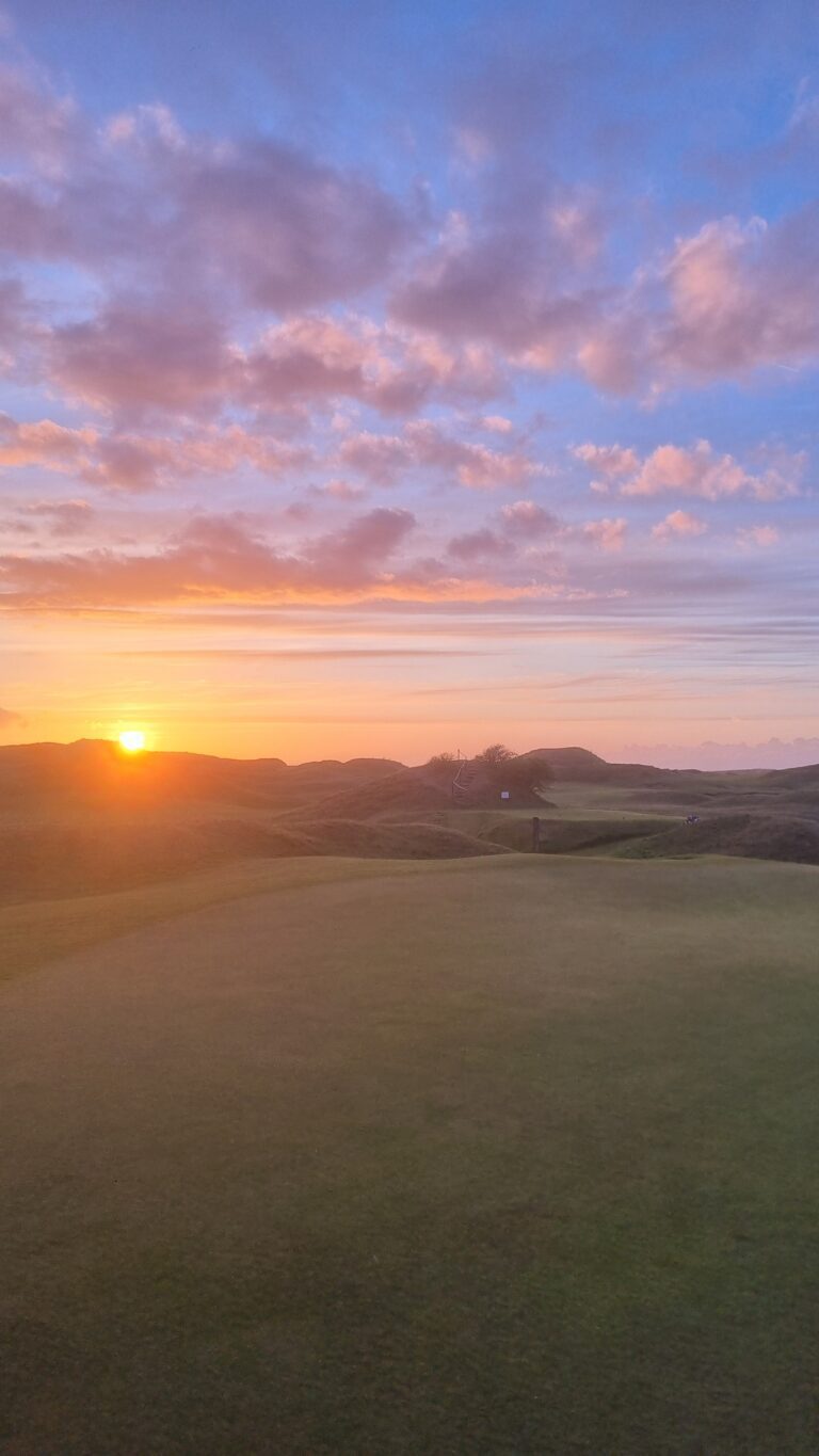 Sunset over rolling grassy dunes with an orange glow and pink-tinged clouds on the horizon.