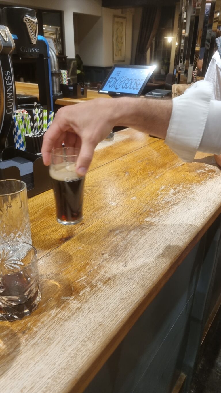 Hand pouring a dark beer into a tall glass at a wooden bar, with Guinness taps in the background.