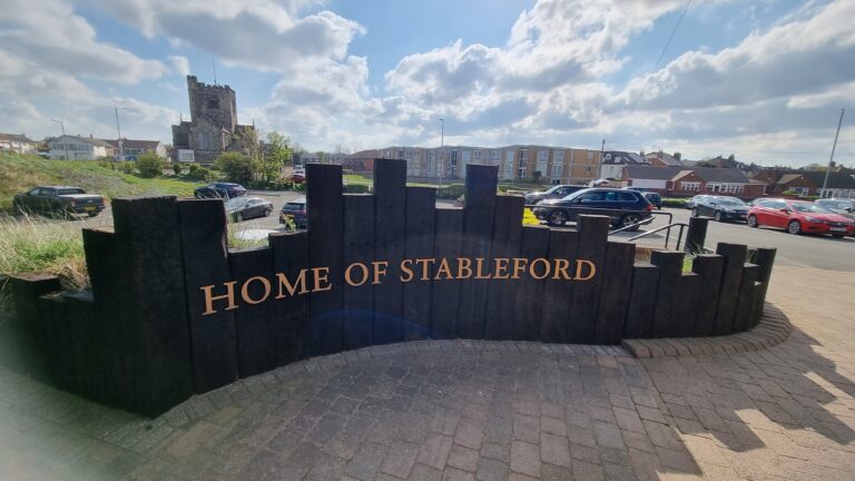 Wooden sign reading 'HOME OF STABLEFORD' in a parking lot with a stone church tower and houses in the background on a sunny day.