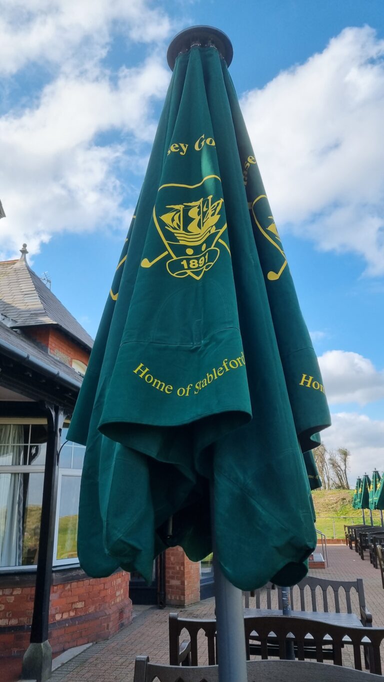 Green outdoor patio umbrella with a yellow crest and text 'Home of Stableford', set against a blue sky and a brick building.
