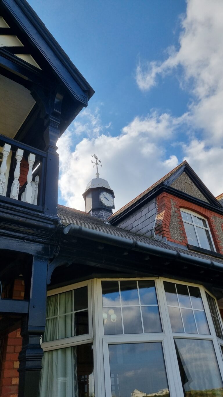 Clock tower with a white clock face on a dark roof, topped by a weather vane, against a blue sky.