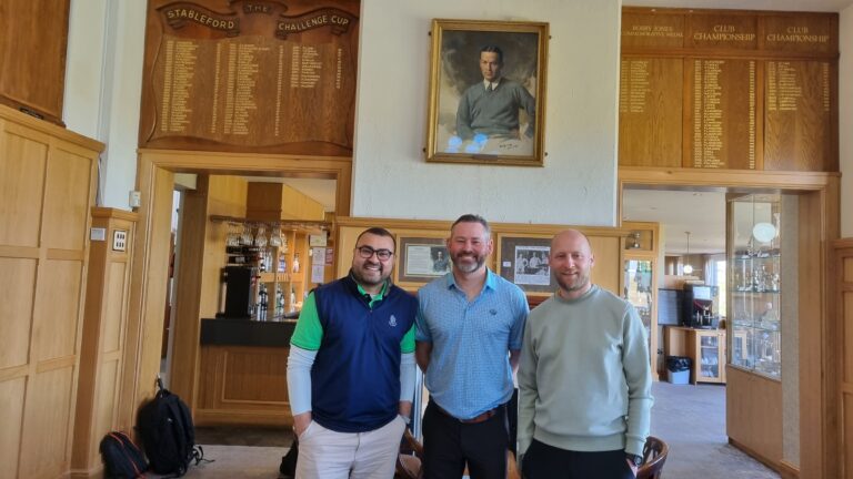 Three men smile for a photo in a wood-paneled club lobby with plaques and a portrait above.