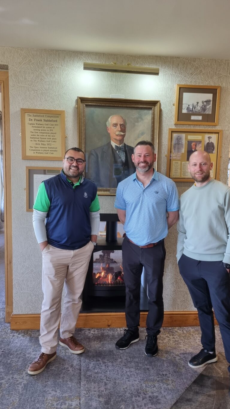 Three men pose indoors in front of a large framed portrait above a lit fireplace with plaques on the wall behind them.