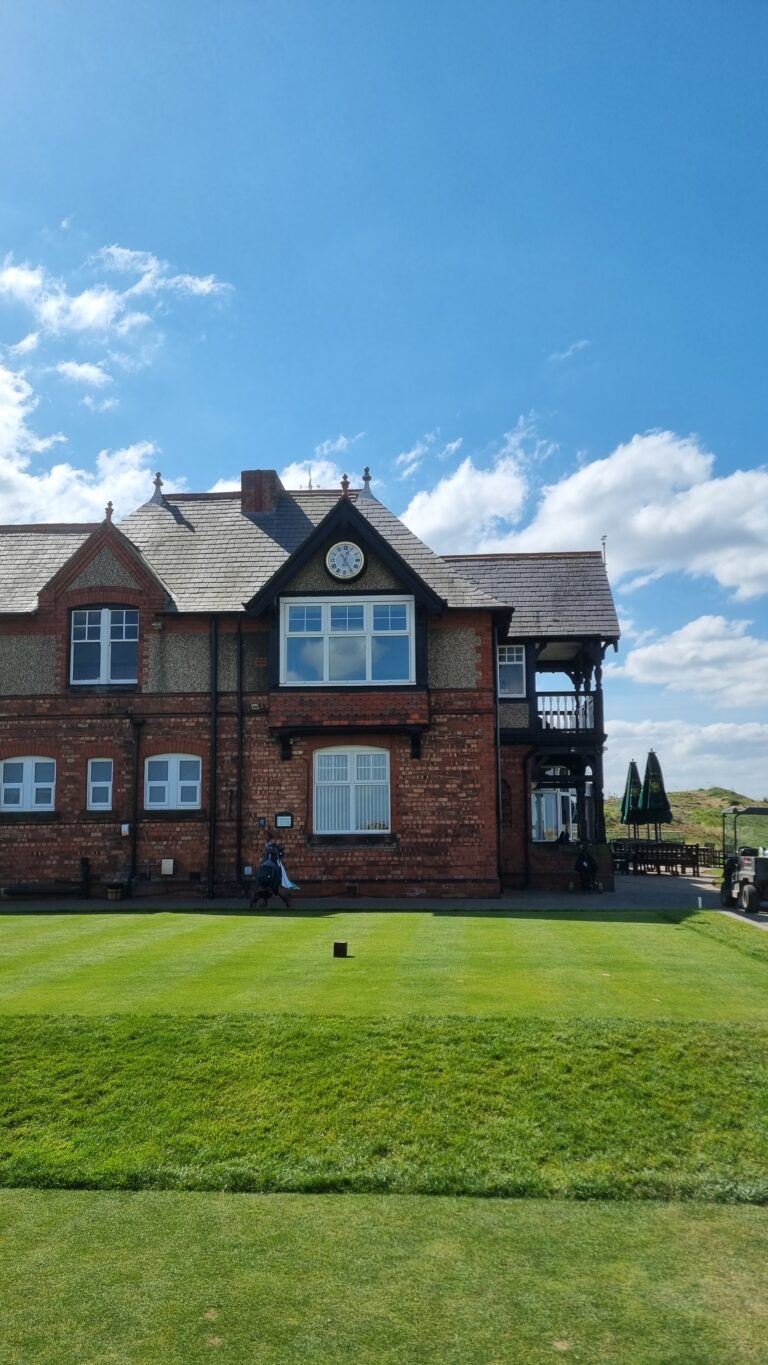 Brick Victorian-style building with a clock on the gable, white-framed windows, and a balcony, set against a blue sky and green lawn.