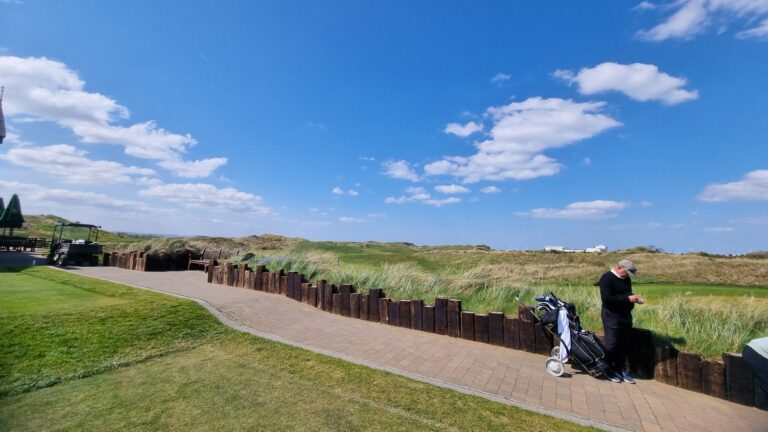 Path along a golf course with a wooden barrier, green grass, and a golfer with a push cart and bag on the right edge.