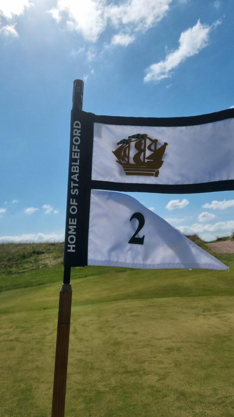 Flag on a golf course reading 'HOME OF STABLEFORD' with a ship emblem and the number 2 against a bright blue sky.
