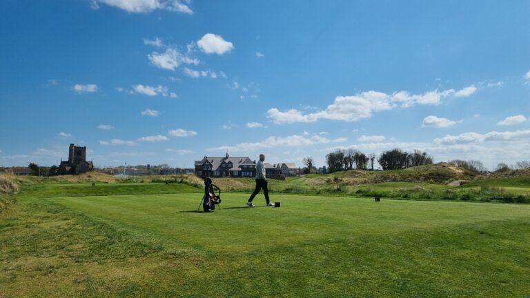 Golfer walking across a green golf course with a bag on a stand and houses in the distance under a blue sky.