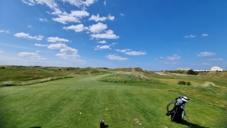 Wide view of a sunny coastal golf course with green fairways, dunes, and a distant white building; a golf bag with clubs is in the foreground on the right.