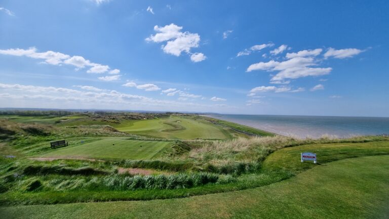 Coastal golf course on a sunny day, with green fairways, dune grasses, and the blue ocean to the right under a clear sky with scattered clouds, a bench in the foreground.