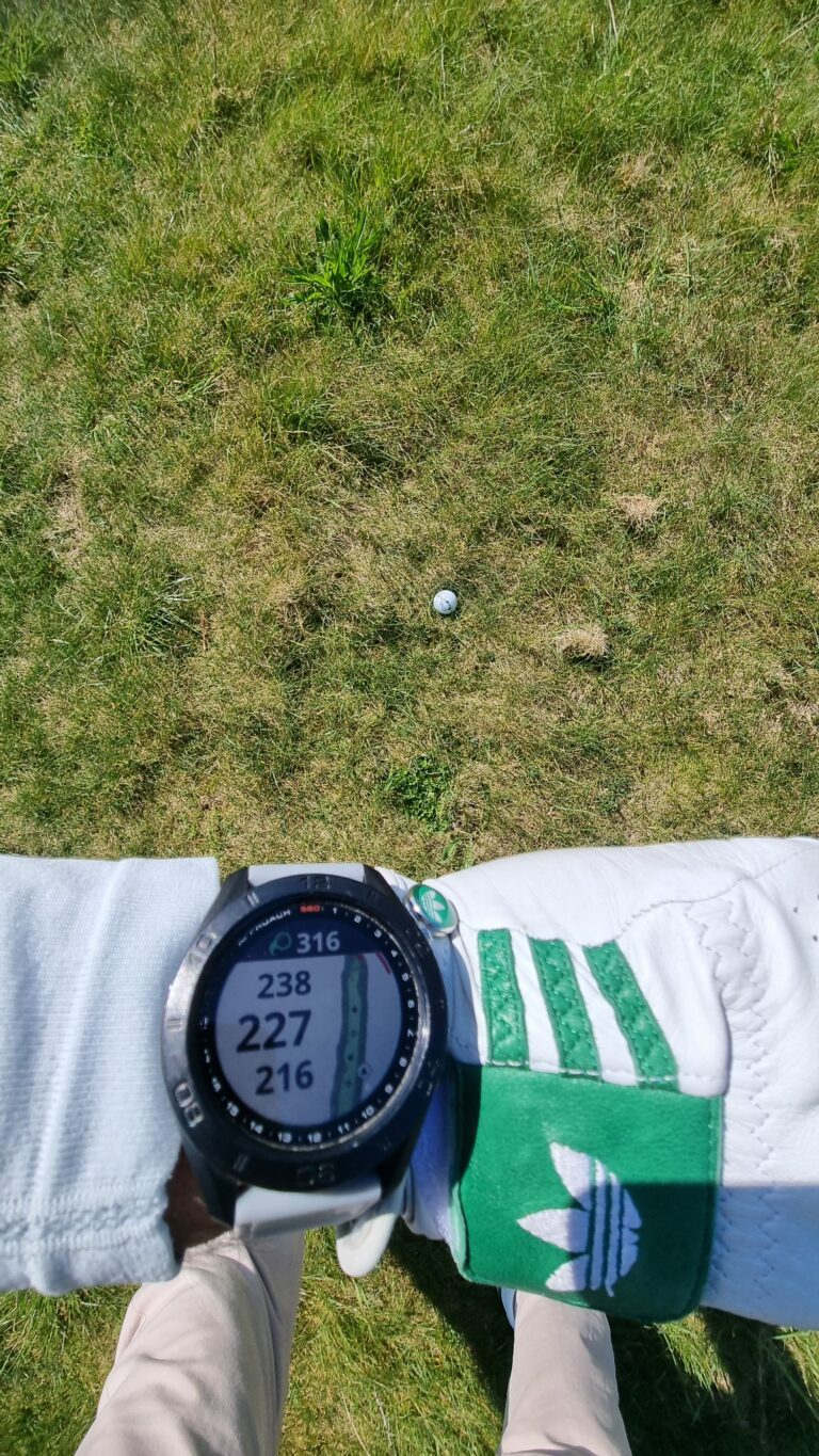 Bird's-eye view of a golfer's gloved arm over grass with a white golf ball near the center and a digital watch visible on the wrist.