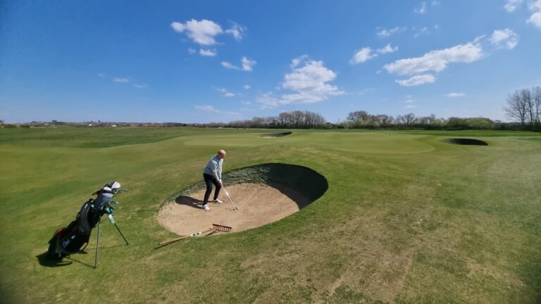 Golfer in a light jacket stands in a sand bunker on a sunny golf course, with a golf bag resting on the grass nearby and clear blue skies above.