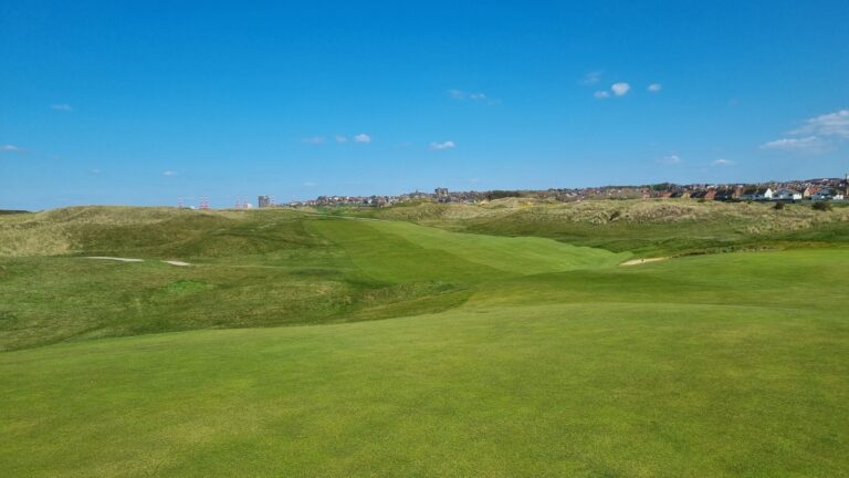 Rolling green golf course with fairways and distant town under a bright blue sky.