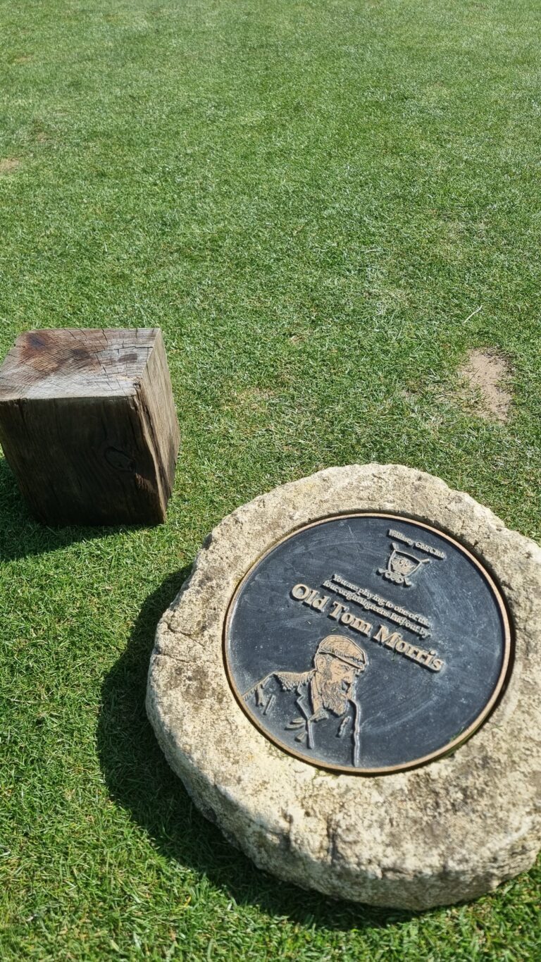Grass field with a large stone pedestal and a round bronze plaque reading 'Old Tom Morris' and showing a man in a hat next to it