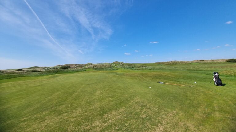 Wide view of a sunny golf course with rolling greens and a distant hill, bag of clubs standing on the right edge.