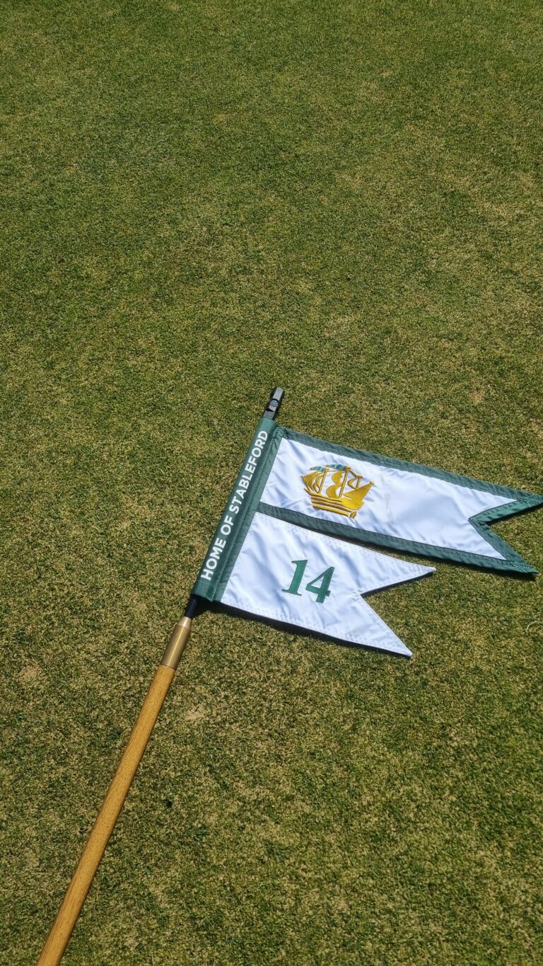Golf flagstick lying on a green putting surface, flag shows hole 14 and 'HOME OF STABLEFORD' text with a crest logo on white and green diagonal flags