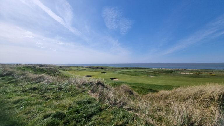 Coastal golf course with green fairways, sand bunkers, and tall dune grass, overlooking the sea under a clear blue sky.