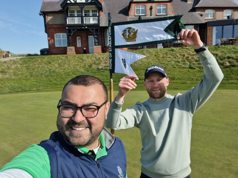 Two men posing for a selfie on a sunny golf course, with a flagstick and flags visible behind them and a large brick clubhouse in the background.