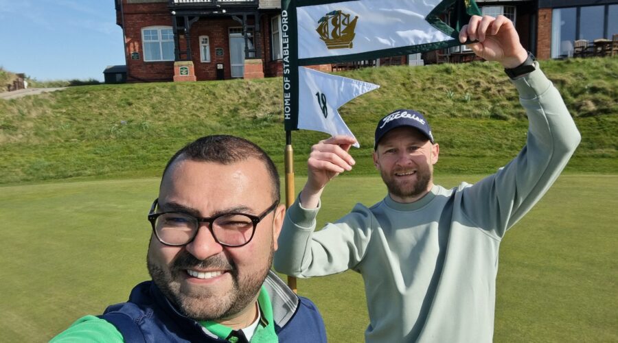 Two men posing for a selfie on a sunny golf course, with a flagstick and flags visible behind them and a large brick clubhouse in the background.