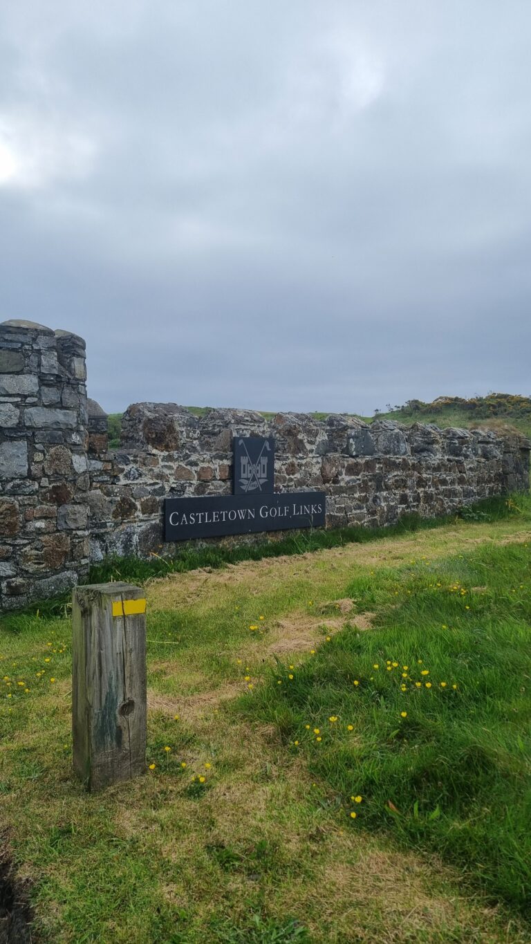 Stone wall with a Castle Town Golf Links sign and logo in a grassy area under a cloudy sky.