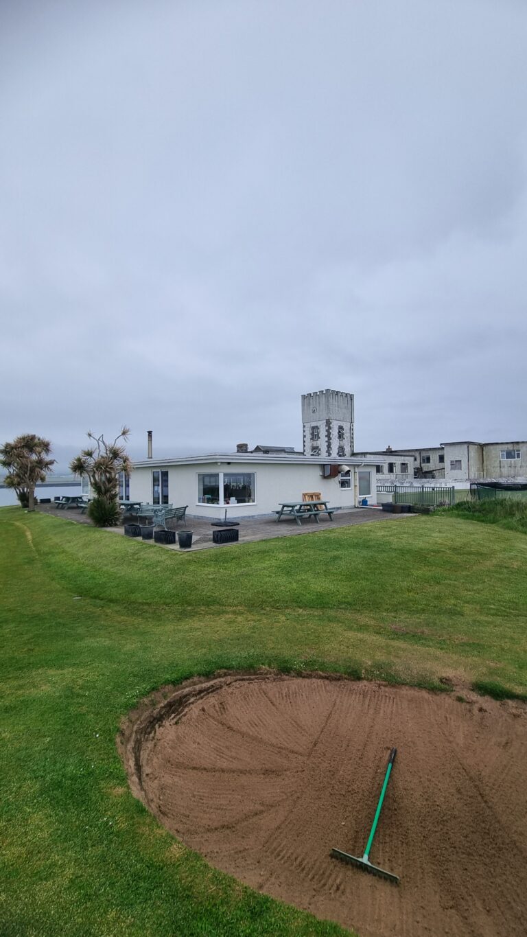 Coastal clubhouse with a white building, outdoor seating, palm trees, and a large sand bunker in the foreground under a cloudy sky.