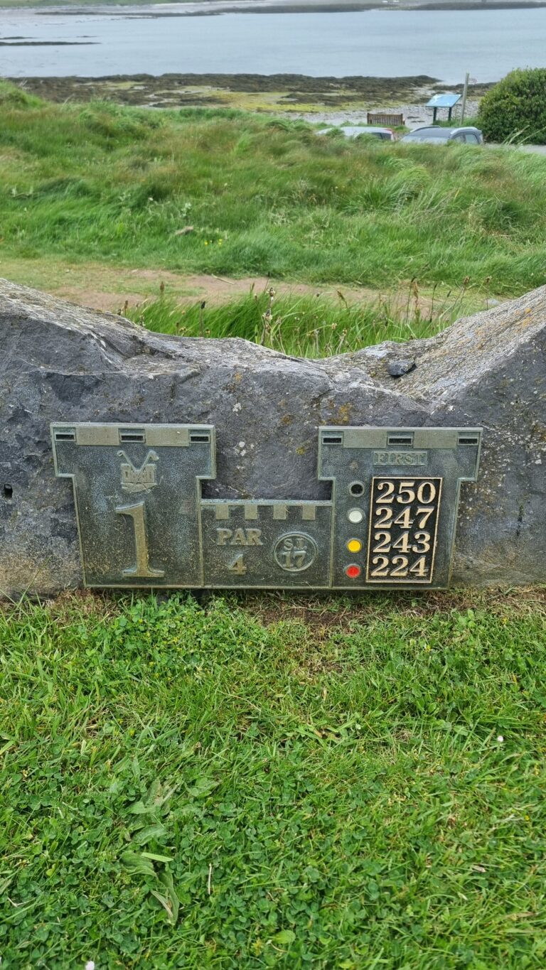 Stone golf-hole marker built into rock wall: hole 1, par 4, with yardages 250, 247, 243, 224 and color indicators, set against a grassy seaside landscape.