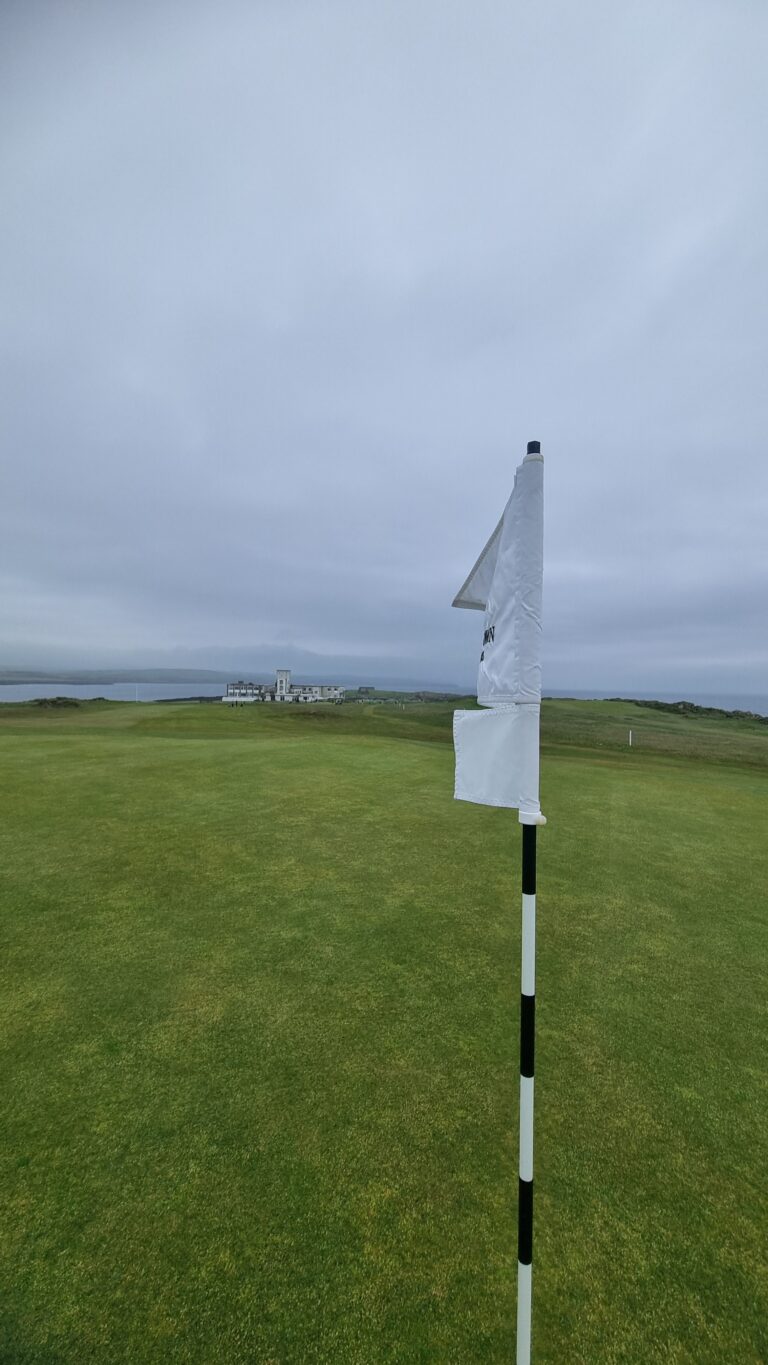 Flag on a white-and-black striped flagstick on a green golf course with the coastline and buildings in the distance under a cloudy sky.