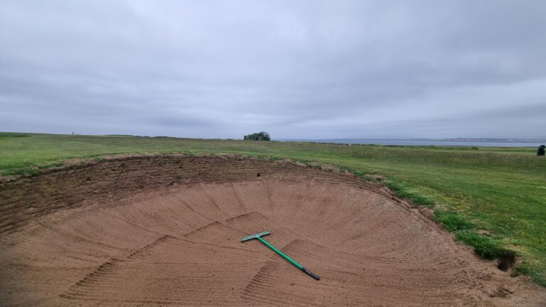 Sand bunker on a grassy golf course with a green rake inside; cloudy sky and distant coastline visible on the horizon.