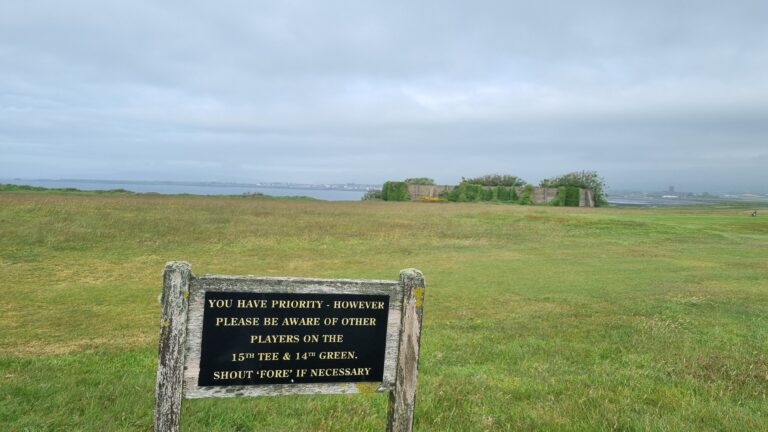 Signboard on a grassy golf course field asking players to yield to others on the 15th tee and 14th green; distant water and hedged wall in background.
