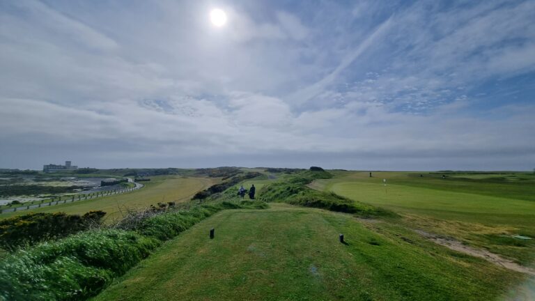 Coastal golf course with a path along the left, green fairways on the right, and a few golfers in the distance under a blue sky, near a building by the shore.