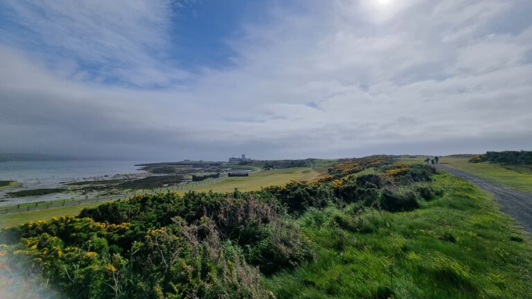 Coastal hillside path along the shore with a distant building, fence, and a few hikers under a cloudy blue sky.