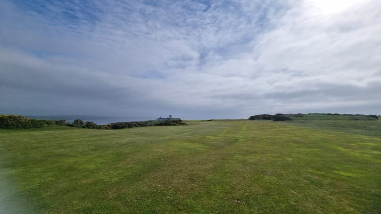 Open grassy field toward the sea with a distant building cluster on the shore under a cloudy sky.