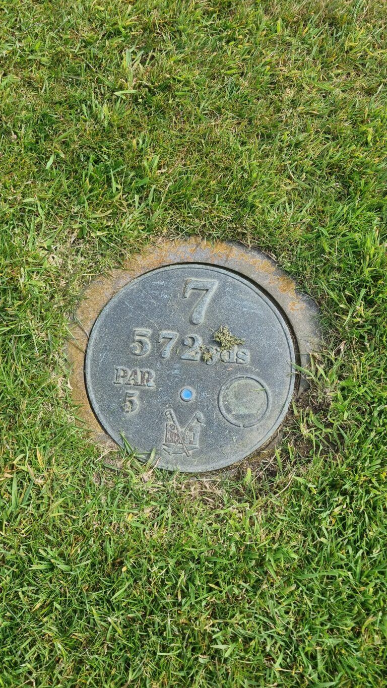 Golf course distance marker on grass reading '7 572 yds PAR 5' with a small blue pin symbol on the cover
